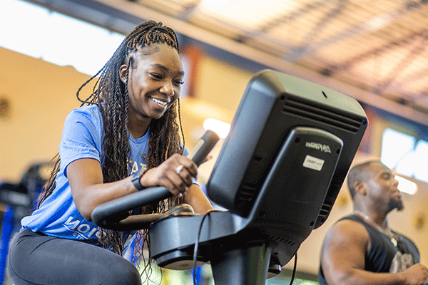 A smiling Black woman with long dreadlocks rides an exercise bike in the Student Recreation Center. She is wearing a light blue t-shirt and dark workout pants. Visible to her right is a smiling, muscular Black man with facial hair and wearing a black tank top. Daylight streams through elevated windows behind them, and yellow walls and a corrugated ceiling are also visible.